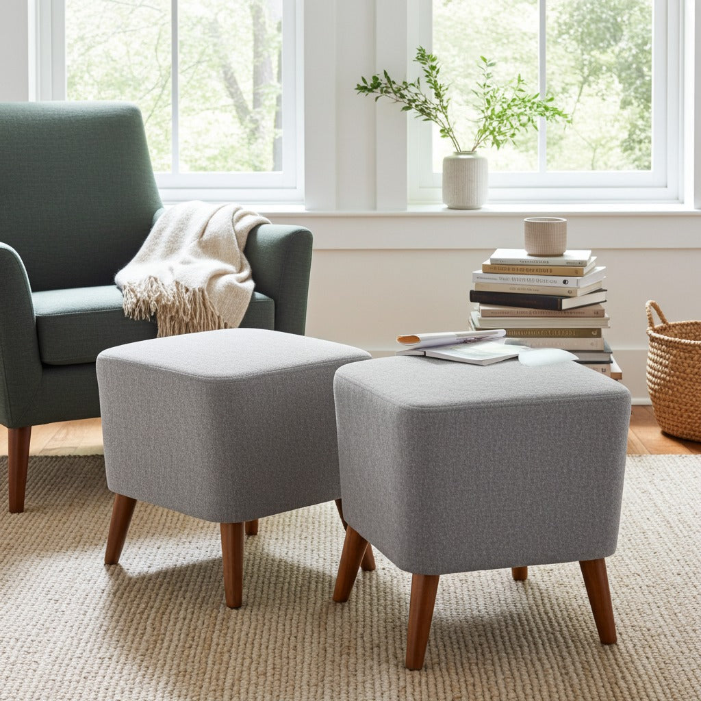Two light gray upholstered ottomans with tapered wooden legs in a bright living room setting, with a window, chair, books, and basket in the background.