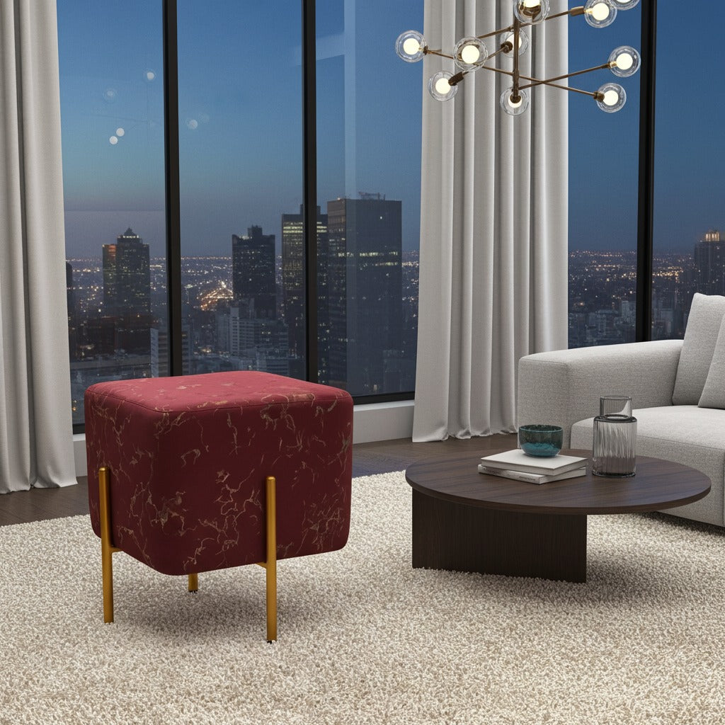 A modern living room scene featuring a square red upholstered ottoman with gold metal legs, positioned next to a dark wood coffee table and a light gray sofa in front of large windows with a city skyline at dusk.