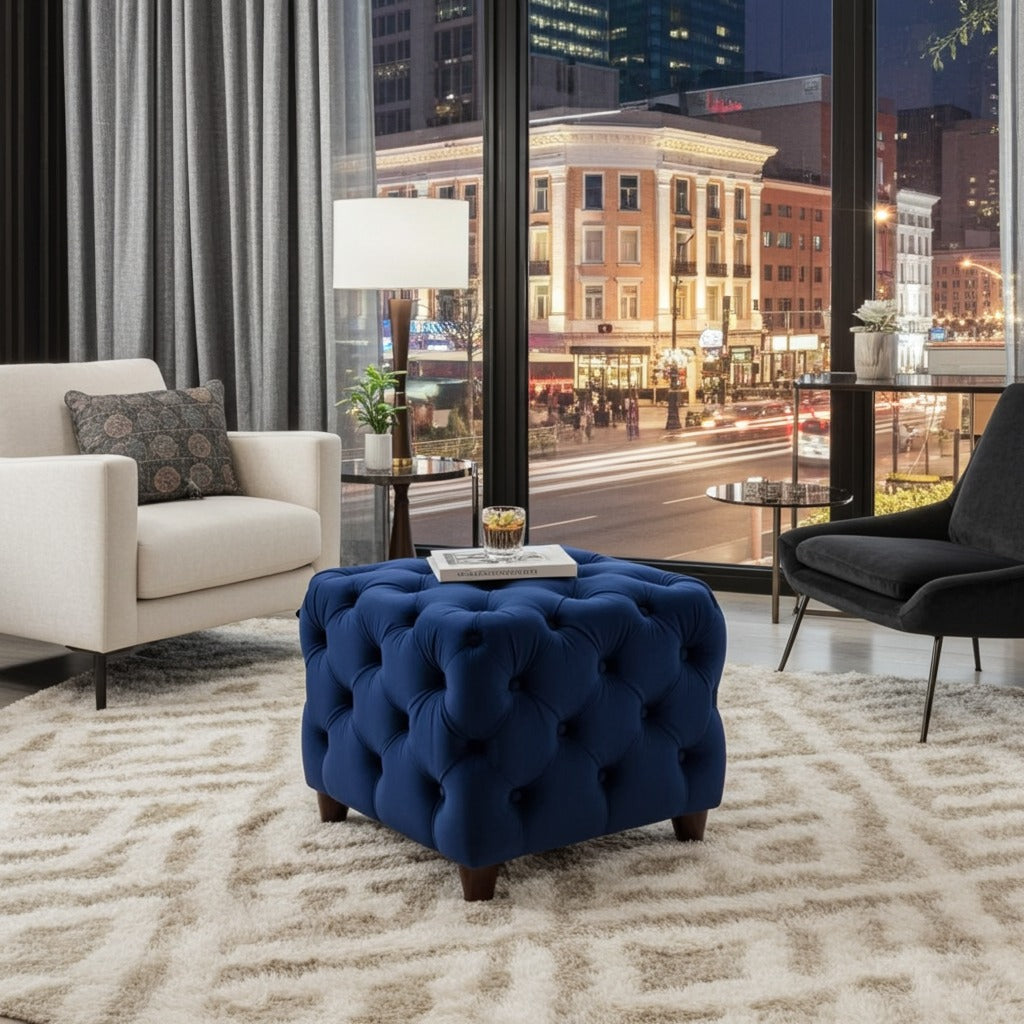 A navy blue tufted velvet ottoman with dark wooden legs sits in a modern living room setting with a white armchair, black accent chair, patterned rug, and a large window showing a city street at night.