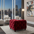 A deep red tufted velvet ottoman with dark wooden legs in a modern living room setting, positioned on a light gray patterned rug in front of large windows showing a city skyline at dusk.