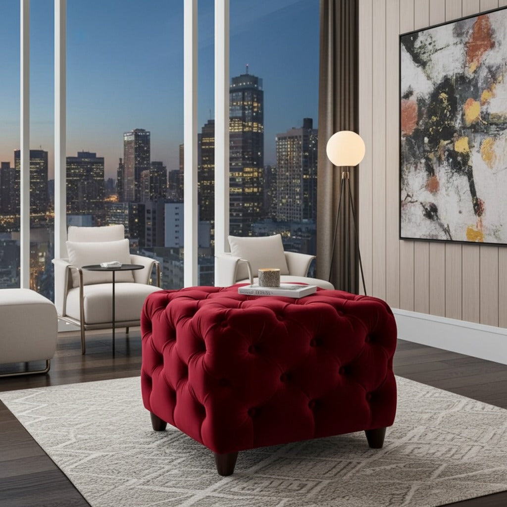 A deep red tufted velvet ottoman with dark wooden legs in a modern living room setting, positioned on a light gray patterned rug in front of large windows showing a city skyline at dusk.