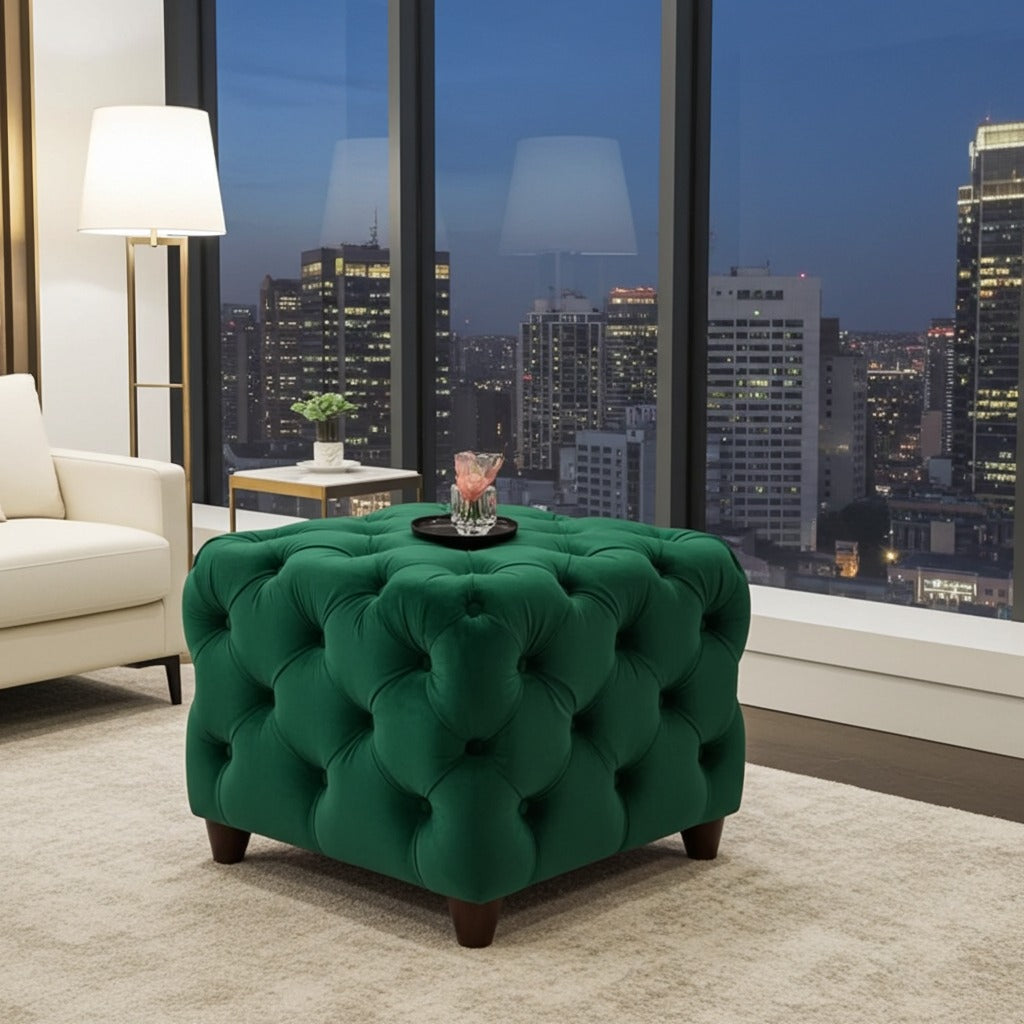 A green tufted velvet ottoman with dark wooden legs sits in a modern living room setting with a city skyline view through large windows in the background.