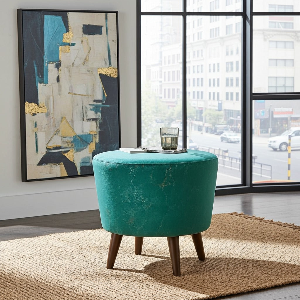 A round teal velvet ottoman stool with four wooden legs sits on a woven jute rug in a modern room setting with a large abstract painting and a window view.