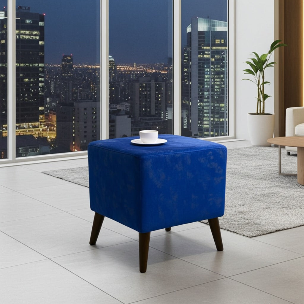 A blue velvet cube ottoman with tapered dark wood legs, styled in a modern living room setting with a white coffee cup on top, set against a large window view of a city skyline at night.