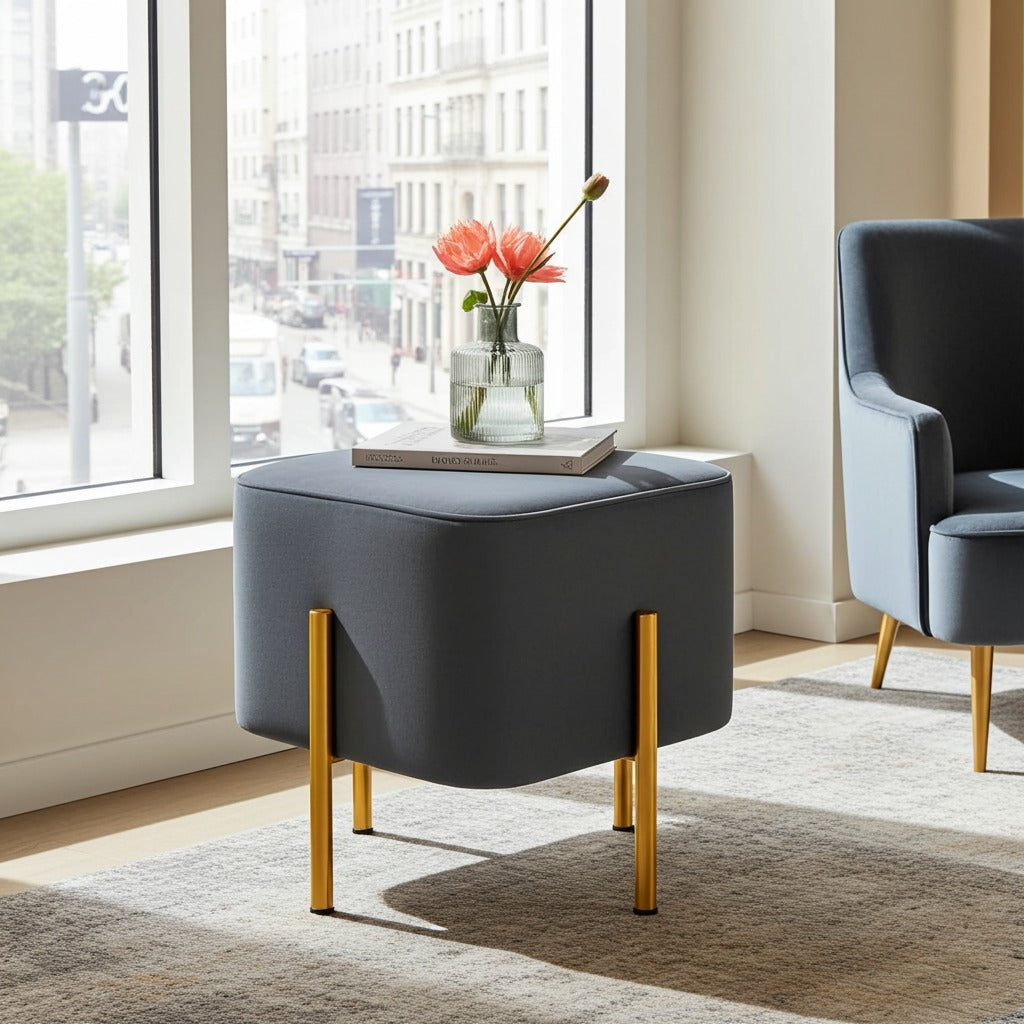 A modern square ottoman stool in dark gray fabric with slim gold metal legs, styled in a bright room next to a window with a vase of flowers on top. A matching gray armchair is partially visible on the right.