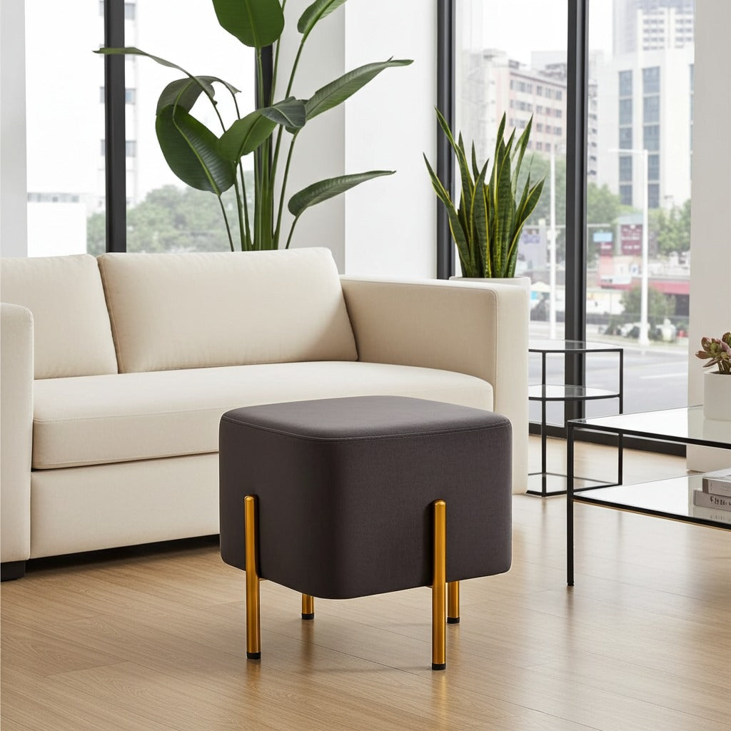 A dark gray square ottoman with gold metal legs, styled in a modern living room setting in front of a cream-colored sofa.