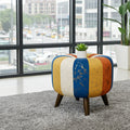 A colorful round pouf ottoman with vertical stripes in blue, yellow, white, and orange, styled on a gray shag rug in front of a large window.