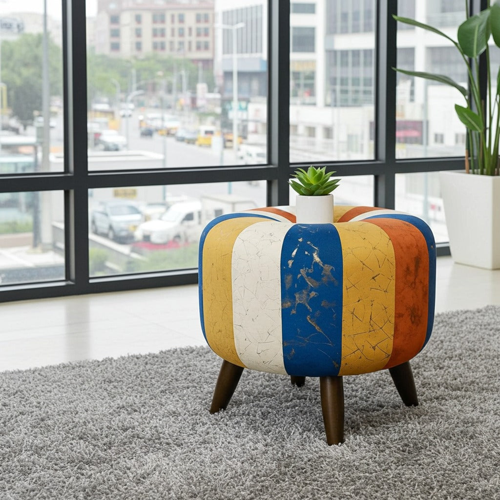 A colorful round pouf ottoman with vertical stripes in blue, yellow, white, and orange, styled on a gray shag rug in front of a large window.