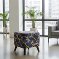 A square upholstered ottoman stool with a geometric pattern in navy blue, yellow, and white, set in a modern living room with large windows, potted plants, and a gray armchair.