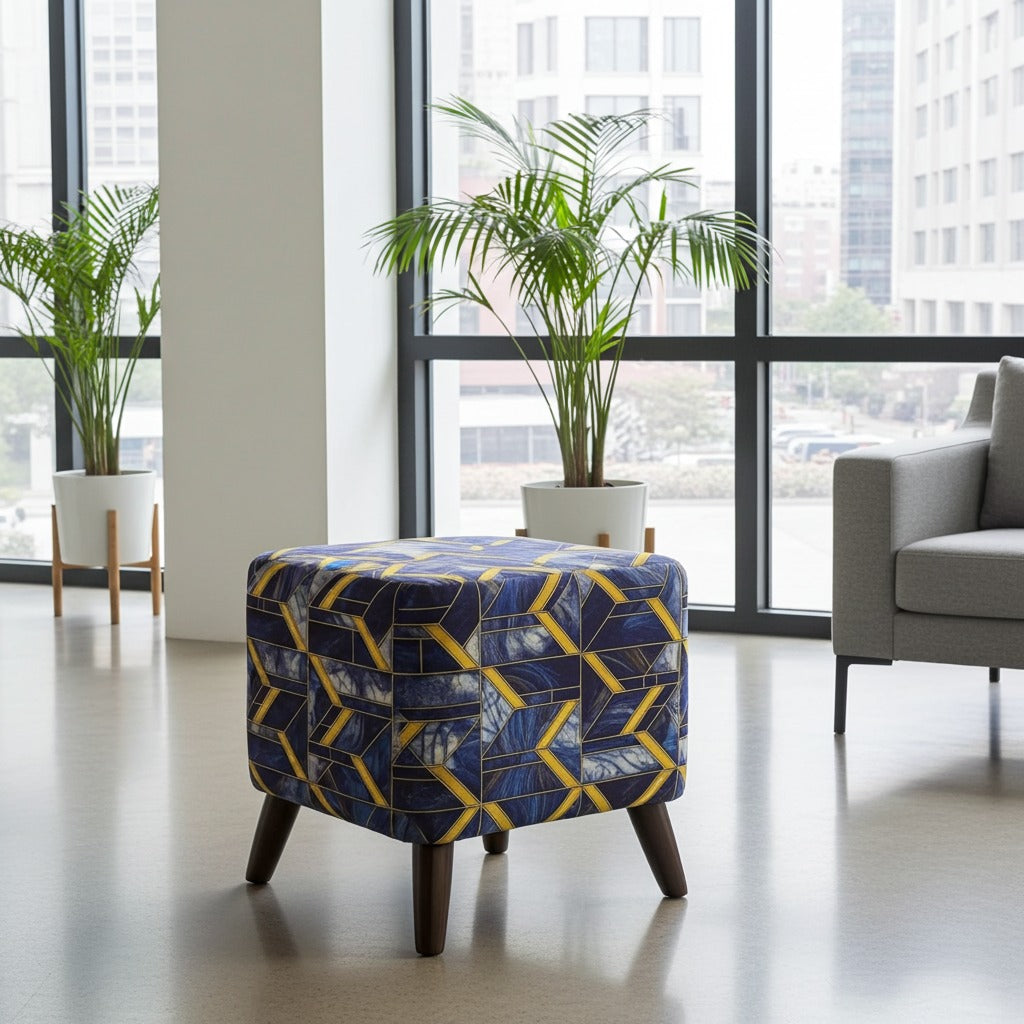 A square upholstered ottoman stool with a geometric pattern in navy blue, yellow, and white, set in a modern living room with large windows, potted plants, and a gray armchair.
