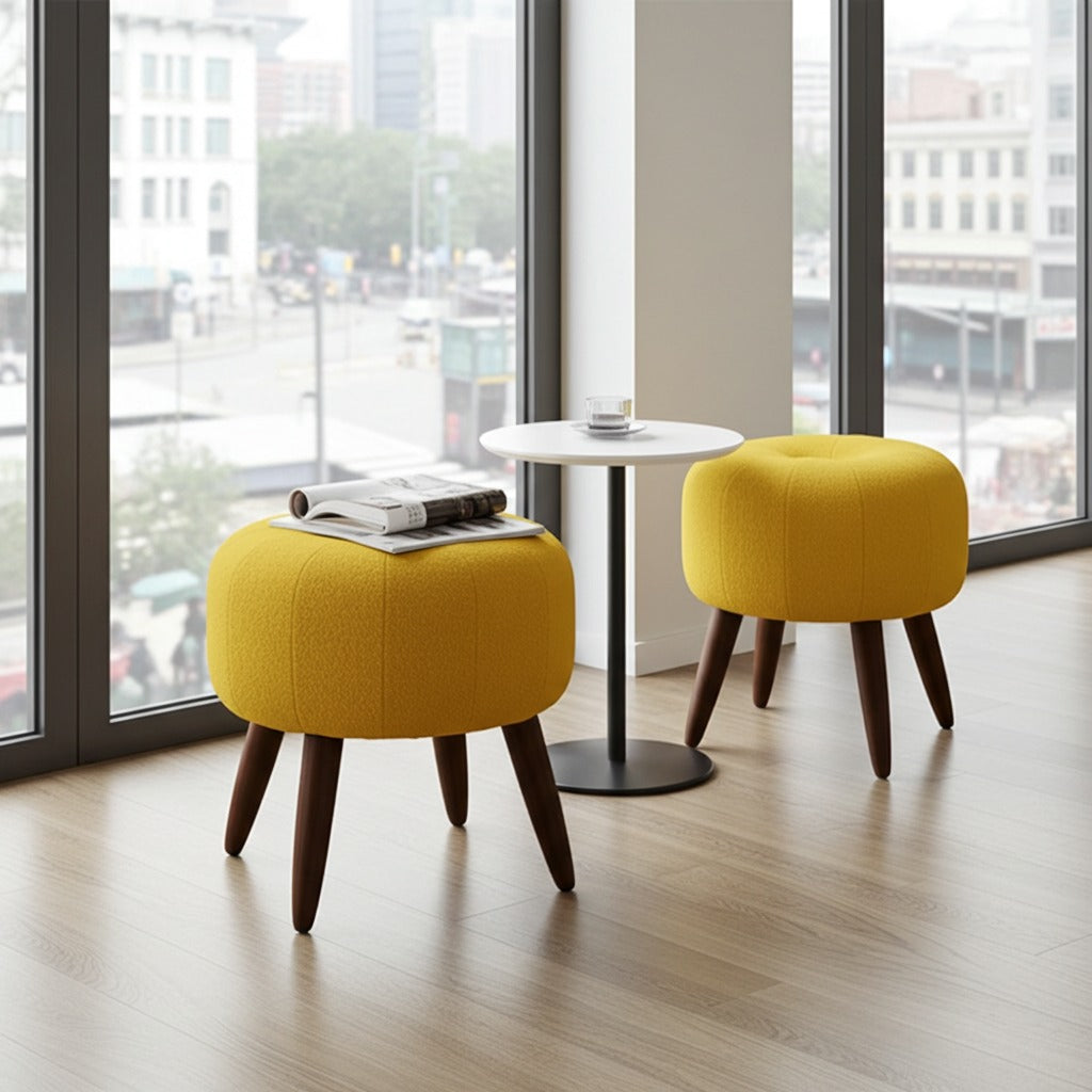 Two round yellow upholstered ottomans with dark wooden legs are shown in a bright room with large windows and a small round table between them.