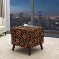 A square ottoman with dark legs, upholstered in a black and brown damask floral pattern, set in a modern living room with a city skyline view in the background.
