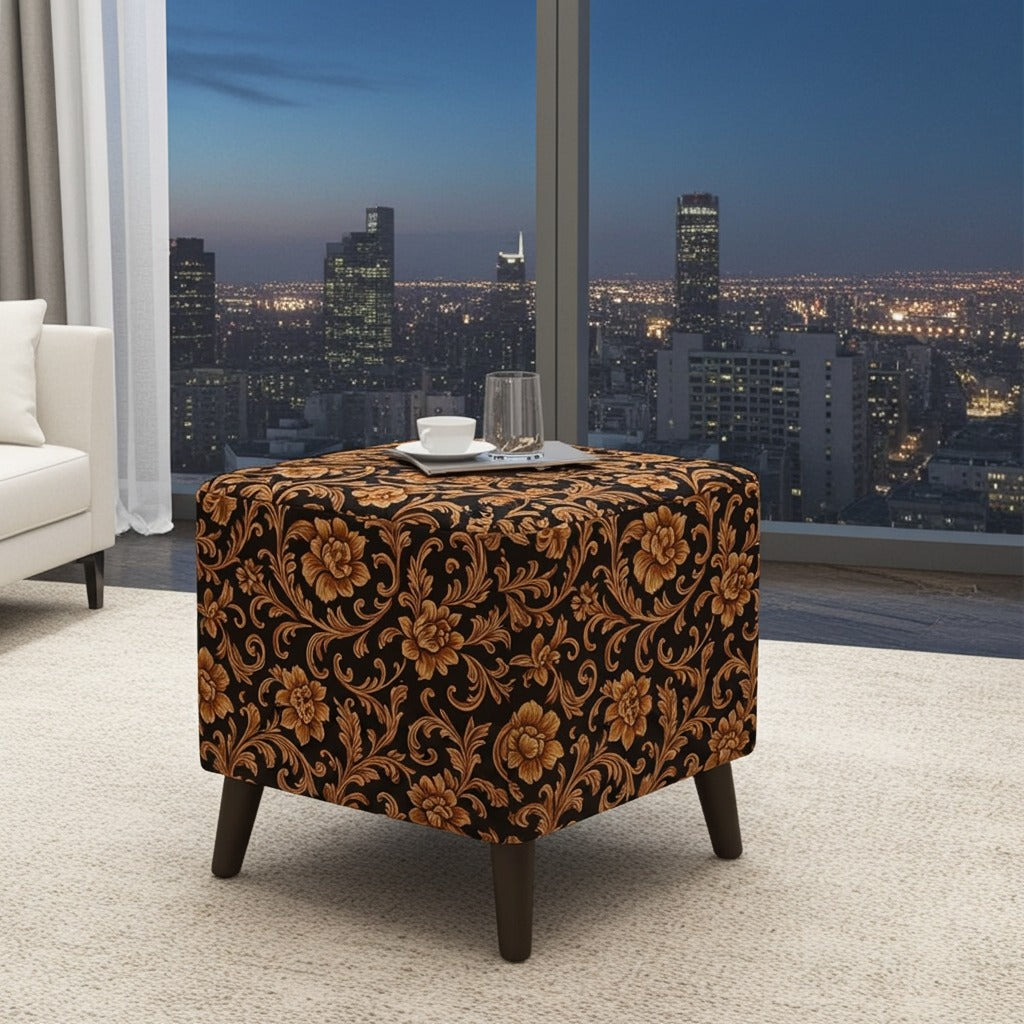 A square ottoman with dark legs, upholstered in a black and brown damask floral pattern, set in a modern living room with a city skyline view in the background.