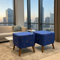 Two square blue ottomans with a gold dragon pattern and wooden legs, placed in a modern living room with a beige sofa and large windows overlooking a city skyline.