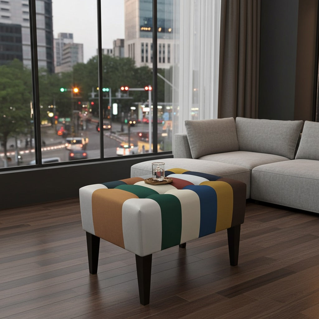 A colorful striped fabric ottoman with dark wooden legs sits in a modern living room in front of a light gray sectional sofa, with a city street scene visible through large floor-to-ceiling windows.
