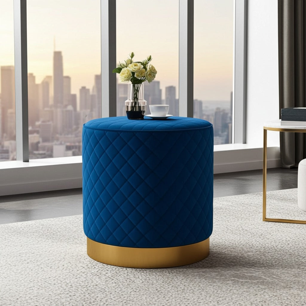 A cylindrical ottoman stool in deep blue quilted velvet with a gold metal base, styled in a modern room with a city skyline view through large windows.