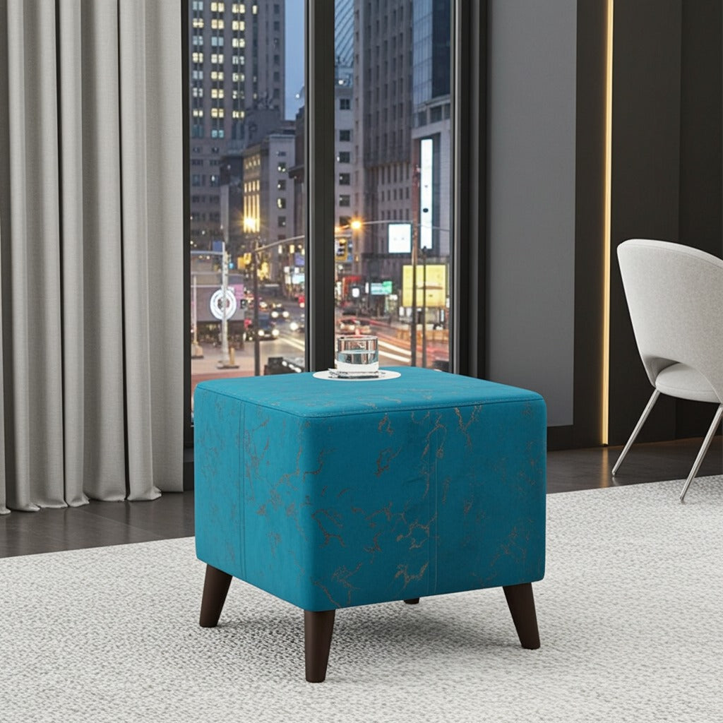 A square ottoman stool in bright turquoise blue fabric with a subtle gold floral pattern and dark brown wooden legs, styled in a modern room setting with a city window view.