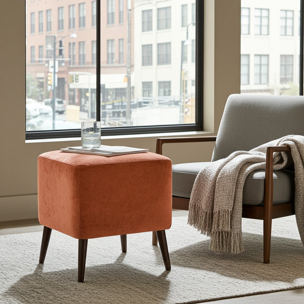 An orange velvet ottoman with dark tapered legs sits on a light rug beside a gray armchair with a beige throw blanket in a bright room with a large window.