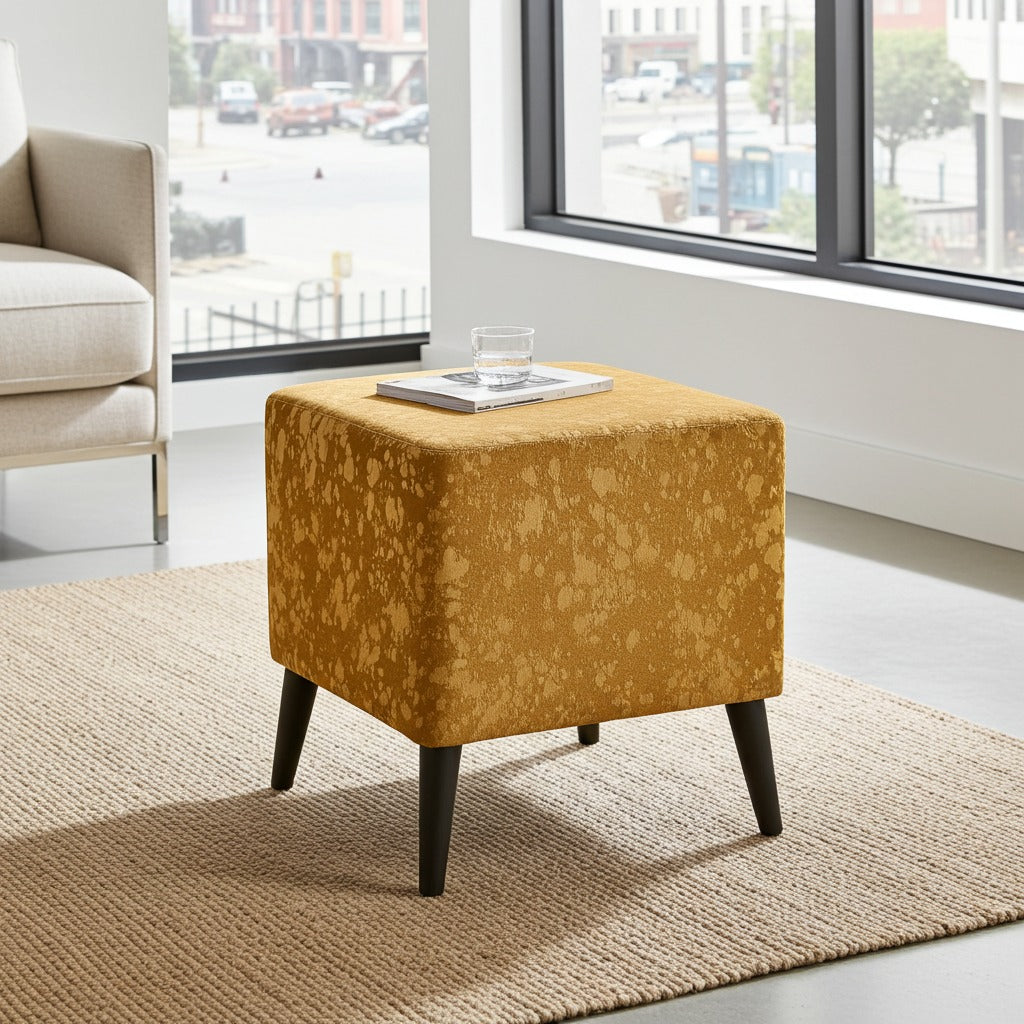 A square upholstered ottoman in mustard yellow fabric with a mottled pattern and dark tapered legs, shown in a bright living room on a woven rug near large windows.