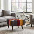 A tufted ottoman with a striped pattern of dark gray, red, and mustard yellow velvet, supported by dark wood legs, styled in a modern living room next to a gray sofa and a glass-top side table.