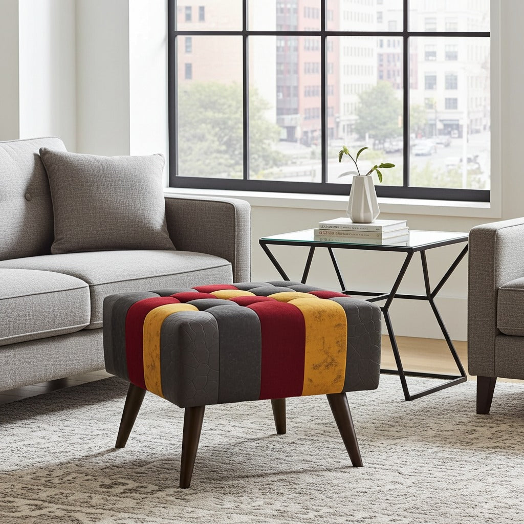 A tufted ottoman with a striped pattern of dark gray, red, and mustard yellow velvet, supported by dark wood legs, styled in a modern living room next to a gray sofa and a glass-top side table.