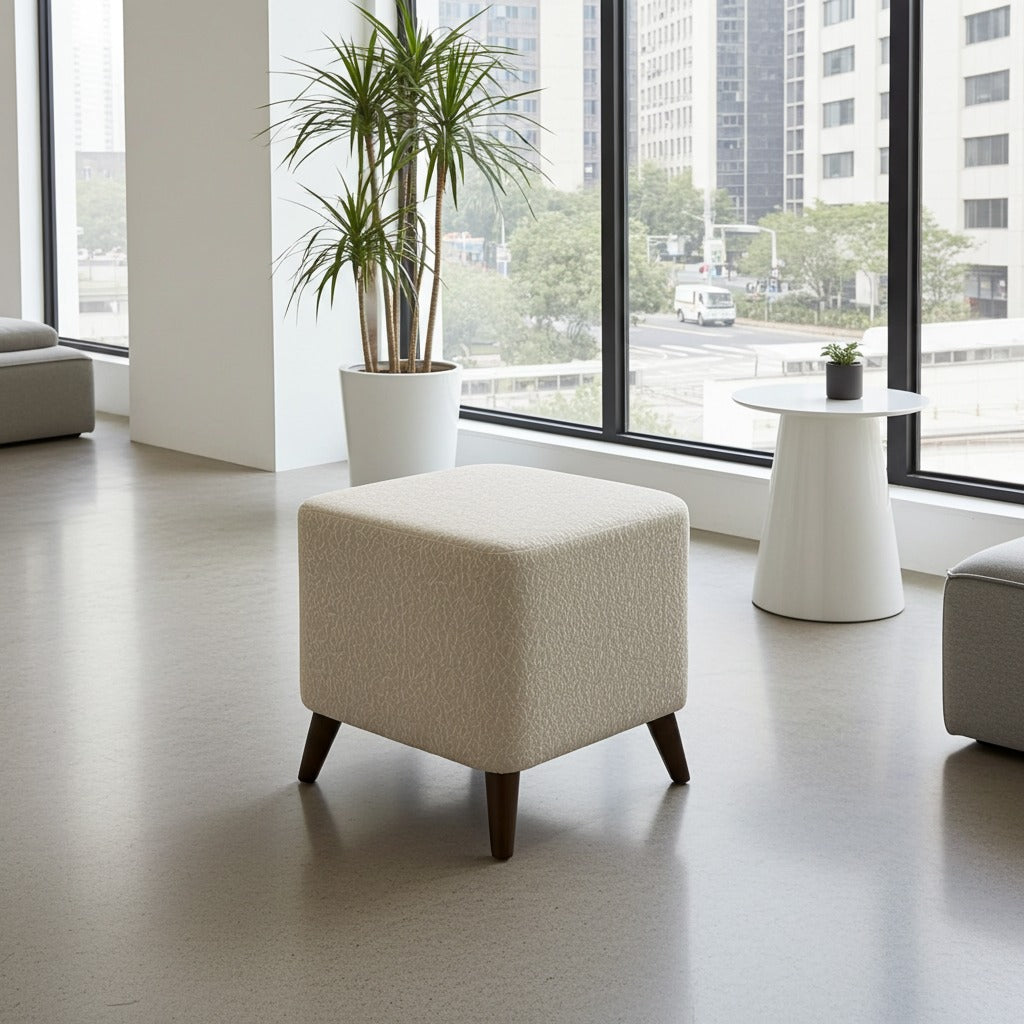 A modern square ottoman with textured cream upholstery and dark wooden legs, positioned in a bright, contemporary room with large windows, a potted plant, and a white side table.