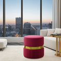 A round upholstered ottoman in bright magenta pink with a metallic gold band around the middle, placed in a modern living room with a white sofa, side table, and large floor-to-ceiling windows overlooking a city skyline at dusk.