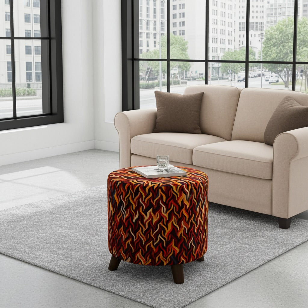A round ottoman with a bold orange, red, and black wavy patterned fabric sits on a gray area rug in a modern living room setting next to a beige sofa.