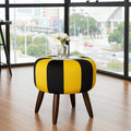 A round upholstered ottoman stool with bold yellow and black vertical stripes, dark brown wooden legs, and a clear glass top holding white flowers, set in a modern room with large windows overlooking a city street.