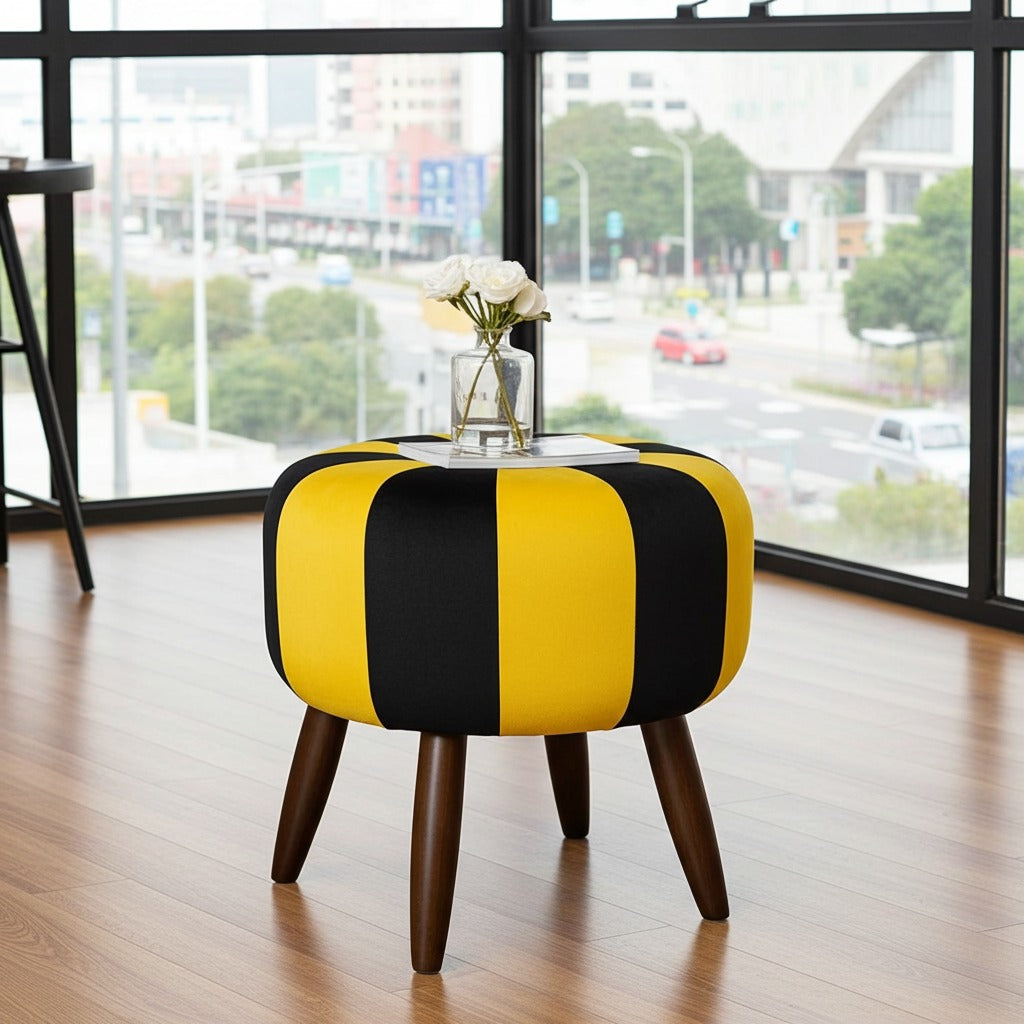 A round upholstered ottoman stool with bold yellow and black vertical stripes, dark brown wooden legs, and a clear glass top holding white flowers, set in a modern room with large windows overlooking a city street.