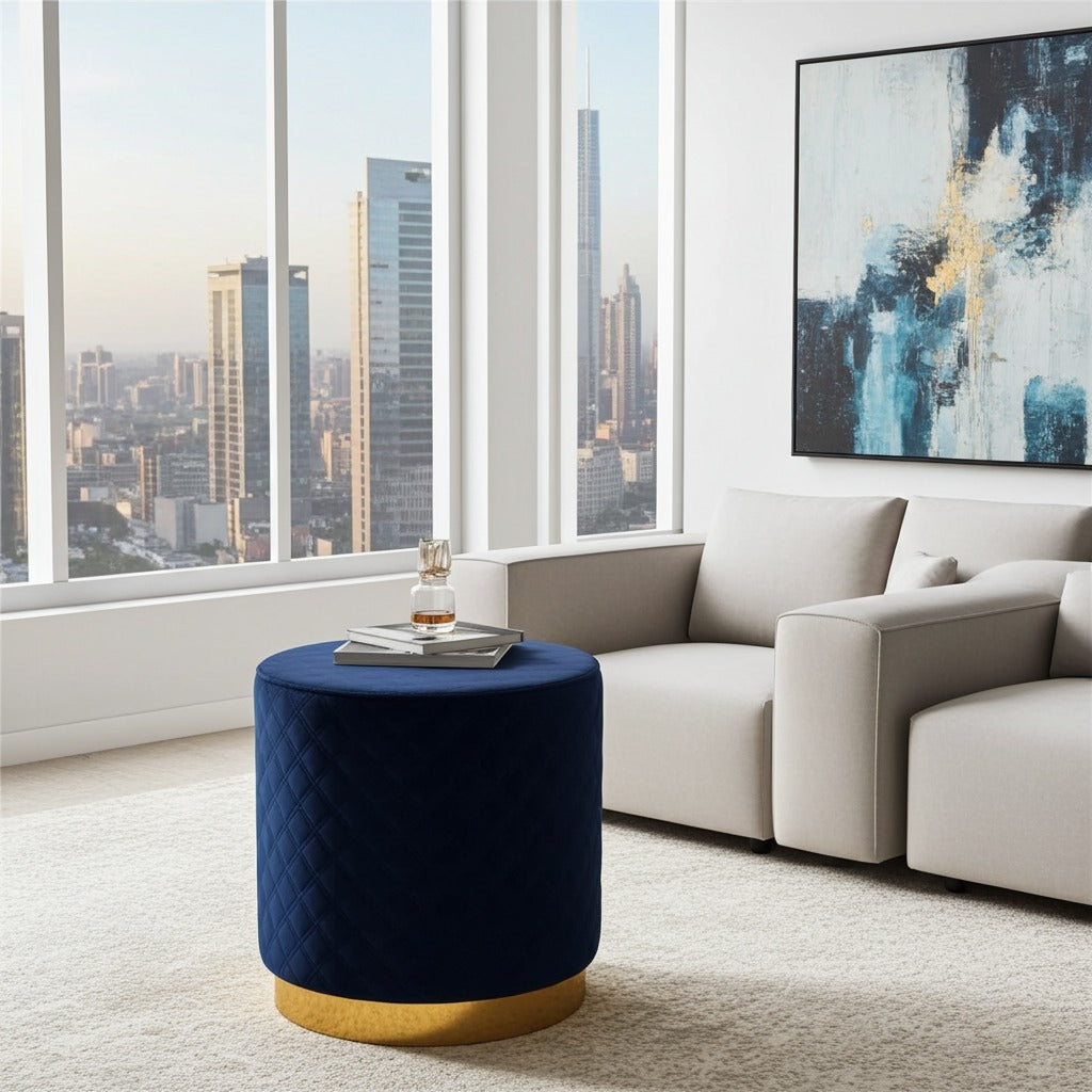 A modern cylindrical ottoman in deep navy blue velvet with a gold base, styled in a bright, contemporary living room with a white sofa and a city skyline view through large windows.