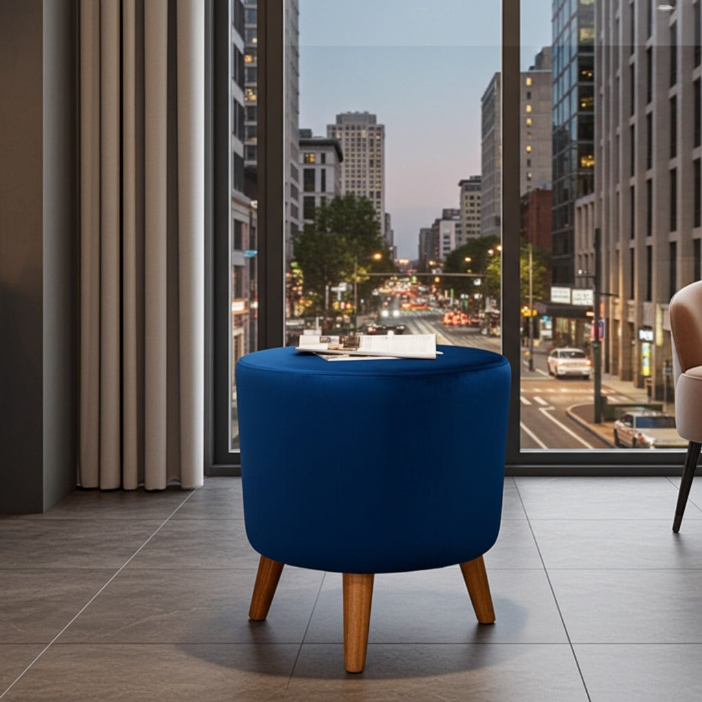 A round blue upholstered ottoman or stool with tapered wooden legs, placed in a modern room with large windows overlooking a city street at dusk.