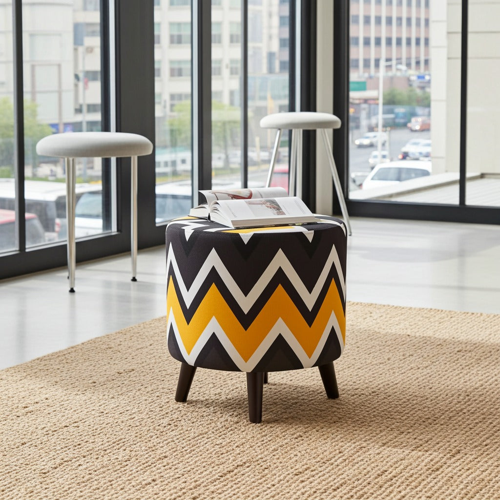 A cylindrical ottoman stool with a bold chevron pattern in black, yellow, and white, supported by four dark wooden legs. It is styled in a modern room setting on a beige rug, with two white stools and a magazine on its top, set against a large window view.