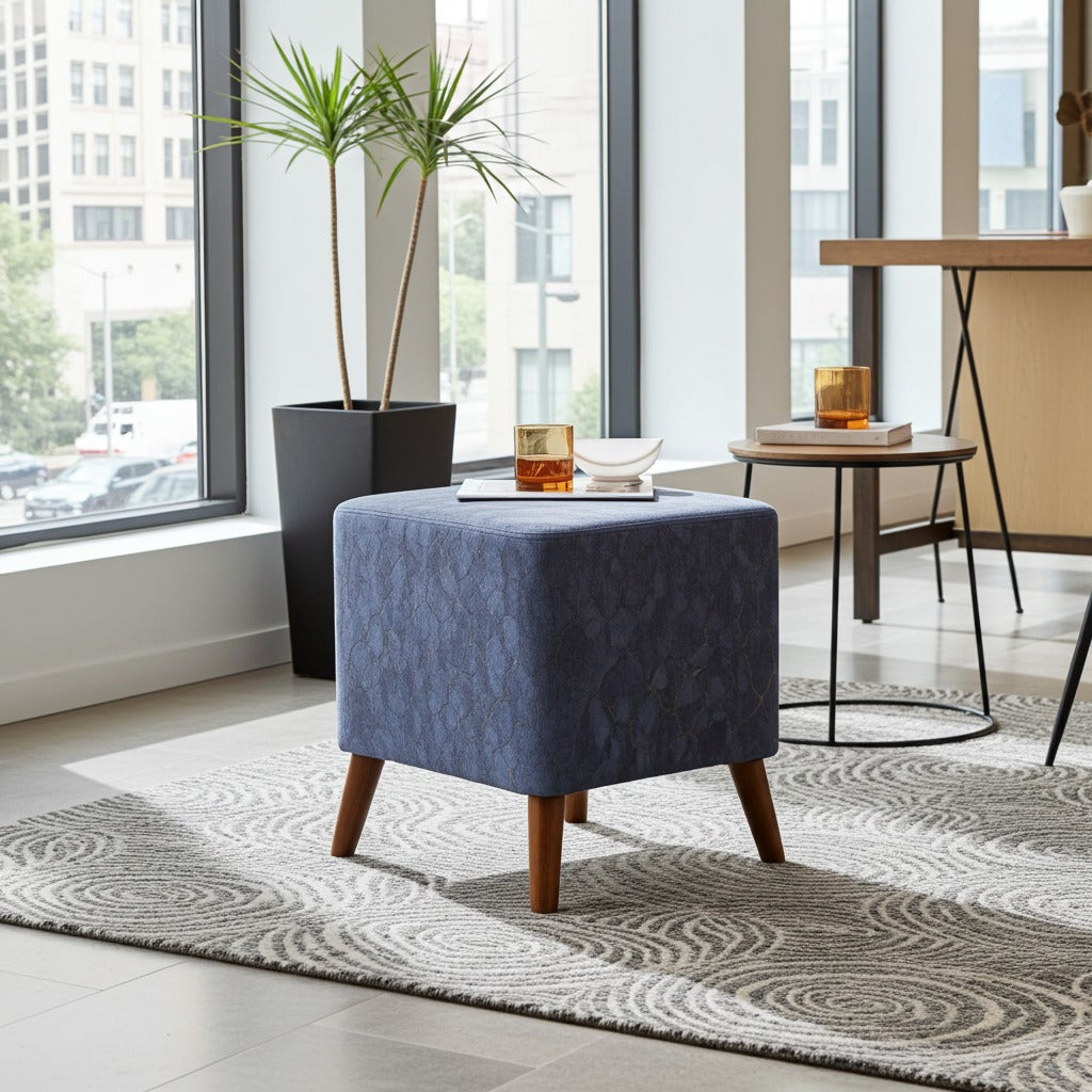 A square upholstered ottoman stool in textured blue fabric with tapered wooden legs, placed on a patterned rug in a bright modern room with large windows, a plant, and side tables.