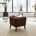 A square brown upholstered ottoman with tapered wooden legs sits on a light beige rug in a bright, modern room with large windows, potted plants, and an open magazine resting on top.