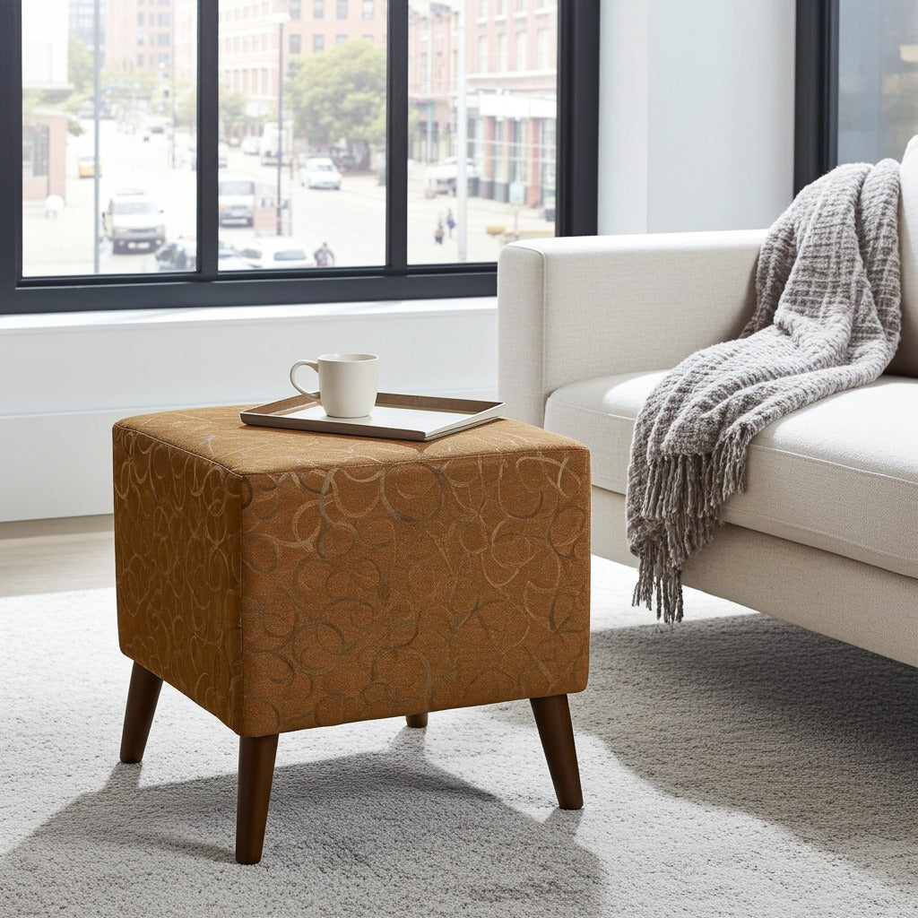 A square ottoman with a textured tan and brown circular pattern and dark wooden legs, styled in a modern living room next to a white sofa with a gray throw blanket.