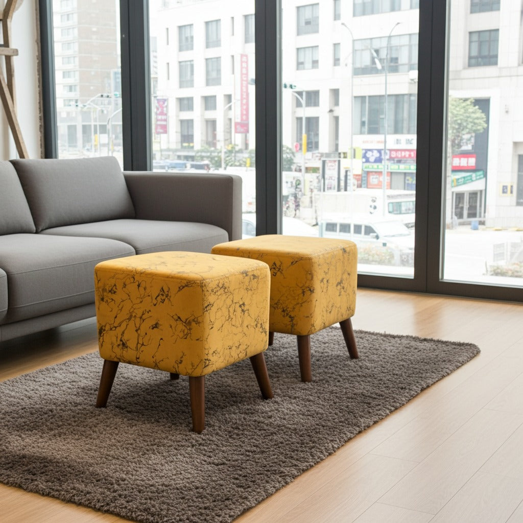Two square ottomans with mustard yellow floral fabric upholstery and dark wooden legs, placed on a gray shag rug in a modern living room with a gray sofa and large windows.