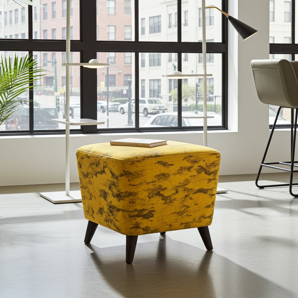 A square yellow upholstered ottoman with a subtle dark abstract pattern and dark tapered legs, styled in a modern room with large windows, floor lamps, and a chair in the background.