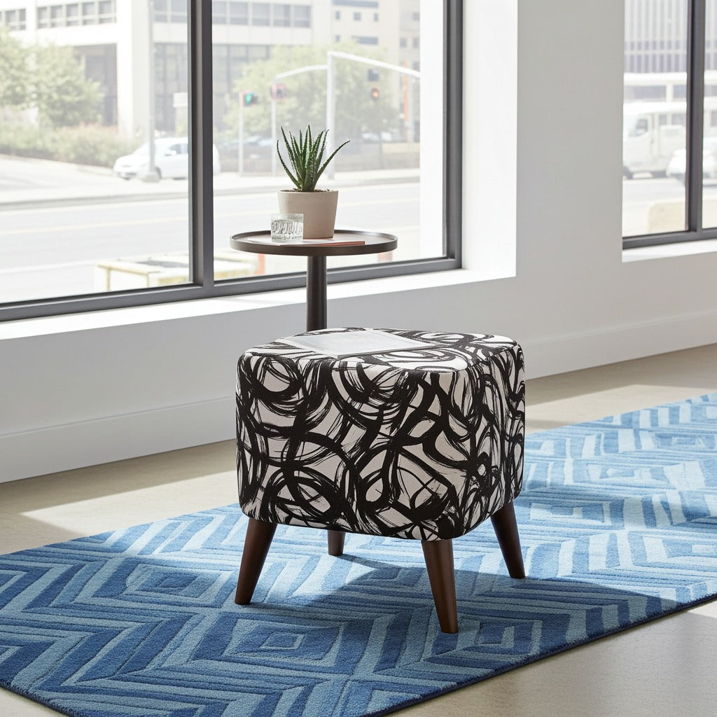 A square upholstered ottoman with a black and white abstract swirl pattern and dark wooden legs, placed on a blue patterned rug in a bright room near large windows.