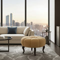 A modern living room scene with a cream sofa, a round tufted ottoman in warm beige, a patterned gray rug, and a large window overlooking a city skyline at sunset.