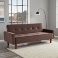 A brown upholstered futon sofa with button tufting on the back and seat, shown in a modern living room setting with a white shag rug, gray walls, a window, and a black floor lamp.