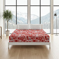 A bed dressed in a red and white retro patterned sheet or duvet, styled in a bright bedroom with a white platform bed frame, white pillows, a potted plant, and a floor lamp, set against large windows showing a mountain view.