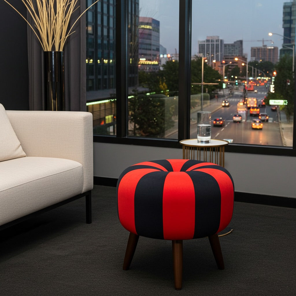 A round upholstered ottoman stool with bold red and black vertical stripes and dark wooden legs, placed in a modern living room setting with a window view of a city street at dusk.
