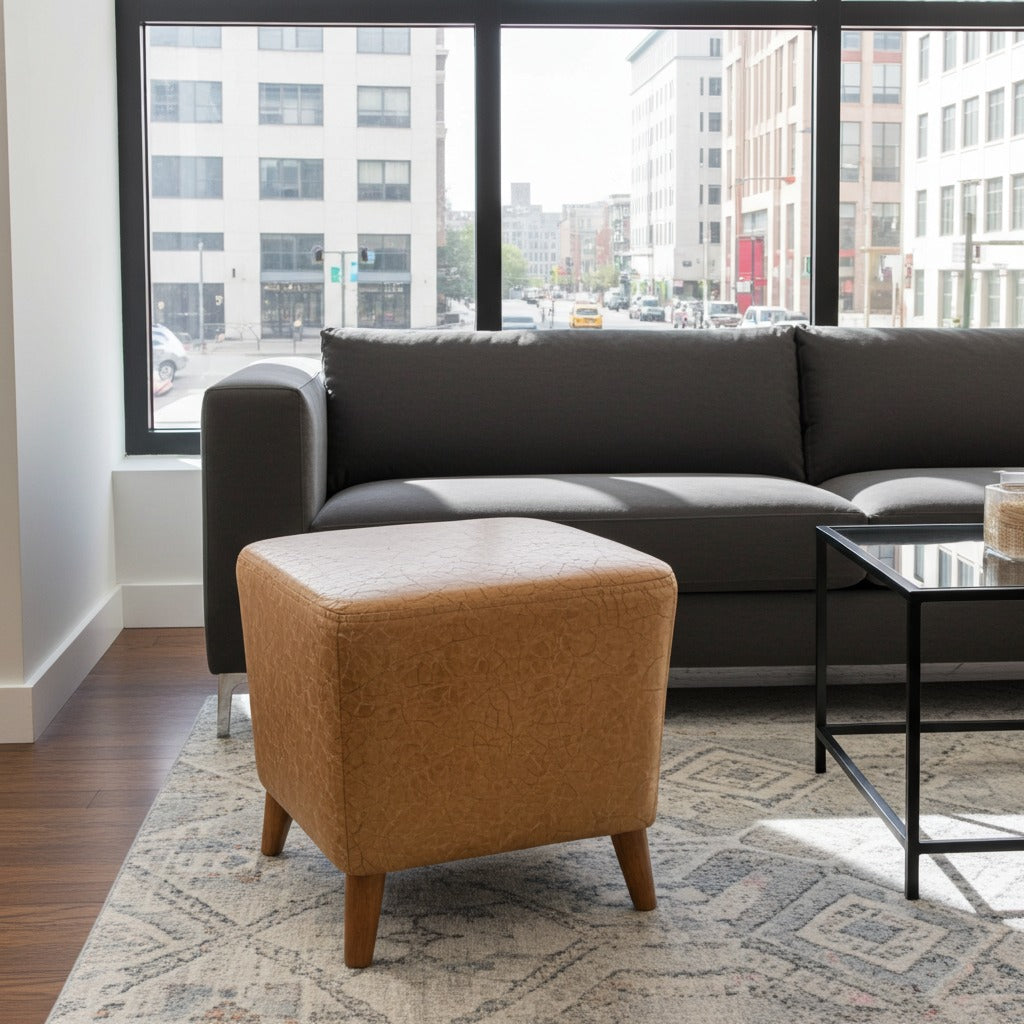A modern living room scene featuring a dark gray fabric sofa and a square tan upholstered ottoman with wooden legs, set on a patterned rug in front of a large window with a city view.