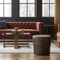 A modern living room scene featuring a rust-brown tufted velvet sofa, a glass-top coffee table with gold metal legs, a round dark brown marble-patterned pouf, and a light gray rug in front of black paneled walls with large multi-pane windows.