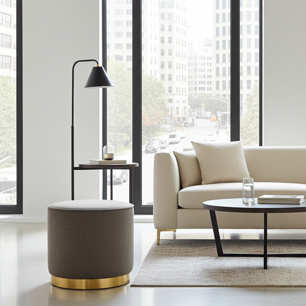 A modern cylindrical ottoman in textured taupe fabric with a polished gold base, styled in a bright, contemporary living room next to a beige sofa and a black floor lamp.