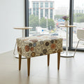 A square upholstered ottoman or bench with a vintage clock face pattern on the seat and four tapered wooden legs, shown in a bright modern room with large windows.