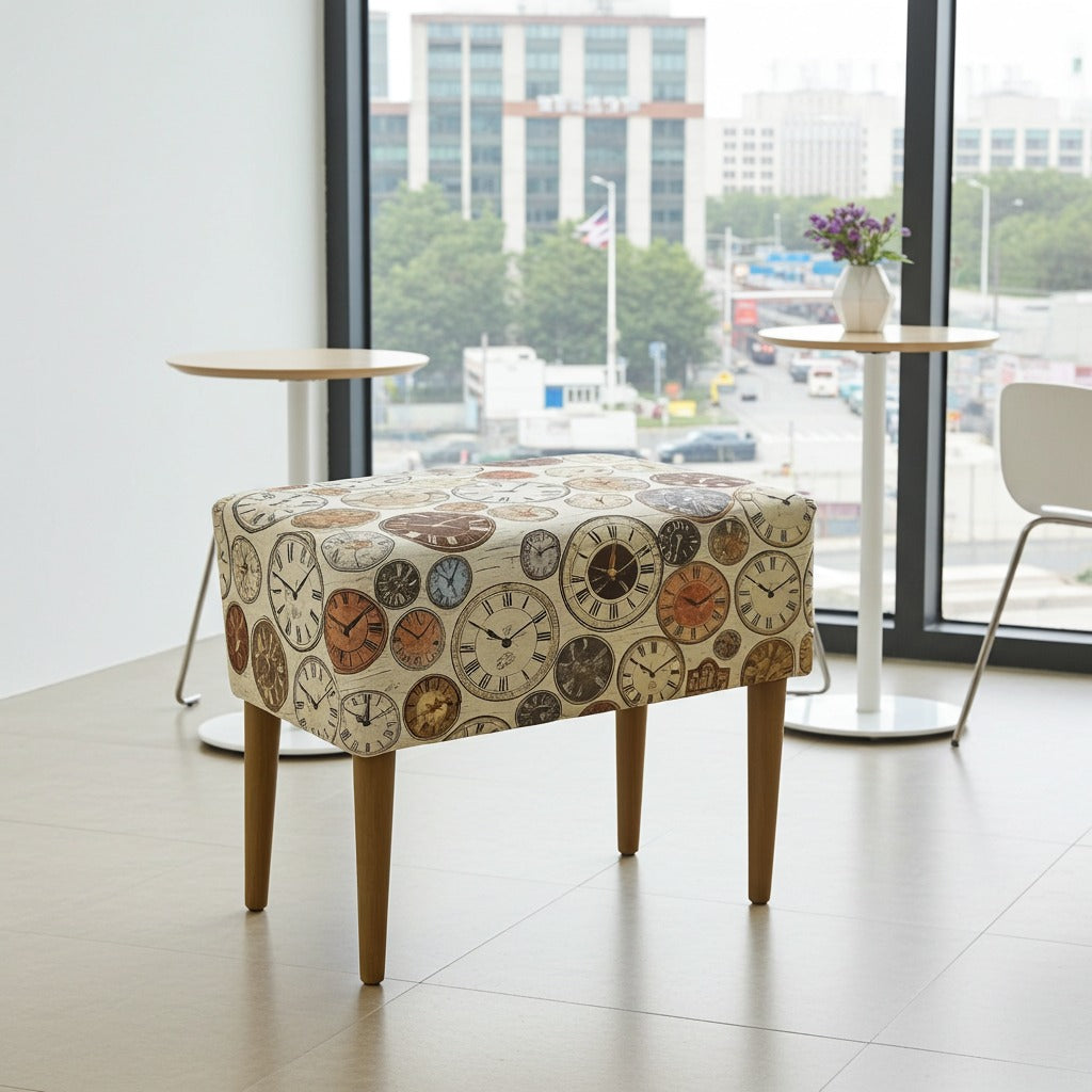 A square upholstered ottoman or bench with a vintage clock face pattern on the seat and four tapered wooden legs, shown in a bright modern room with large windows.