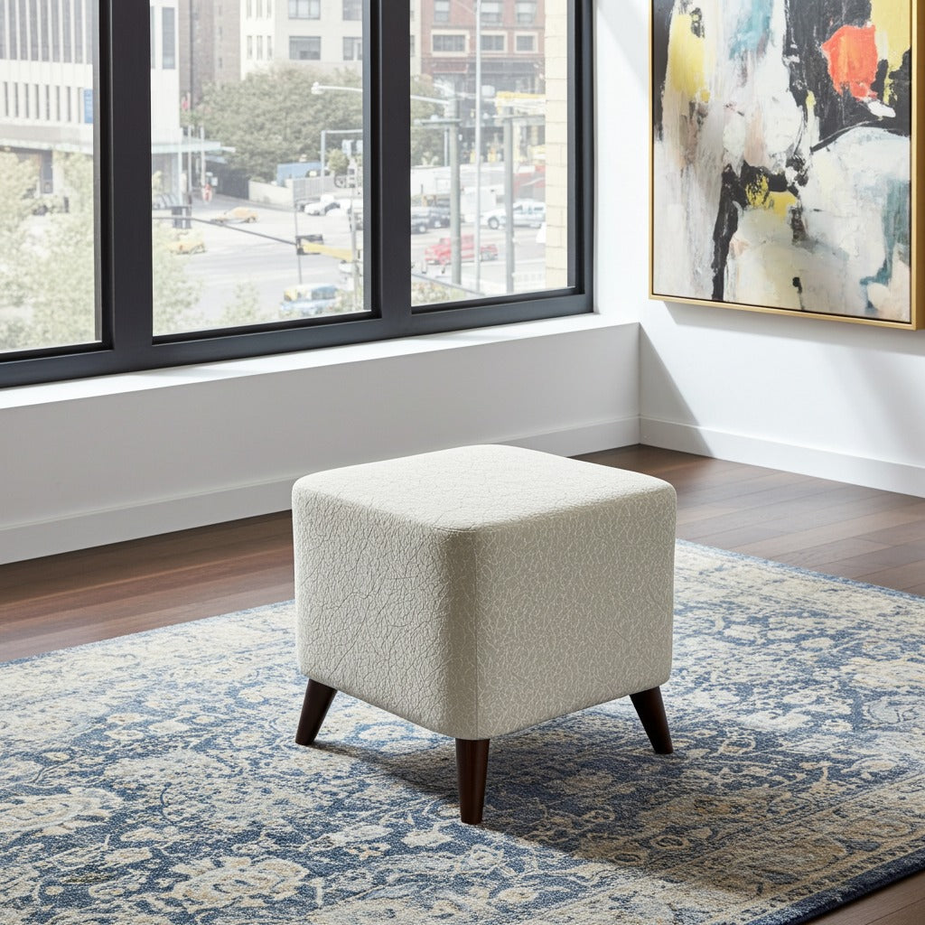 A square upholstered ottoman stool with a textured cream bouclé fabric and dark wooden legs, positioned on a blue patterned area rug in a modern room setting.