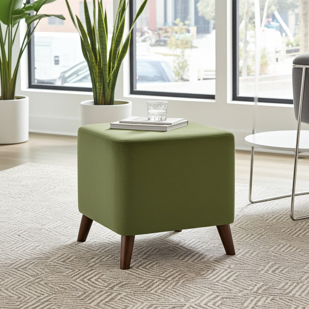 A square ottoman stool in olive green fabric with tapered dark wood legs, styled in a bright living room setting on a patterned rug near large windows.
